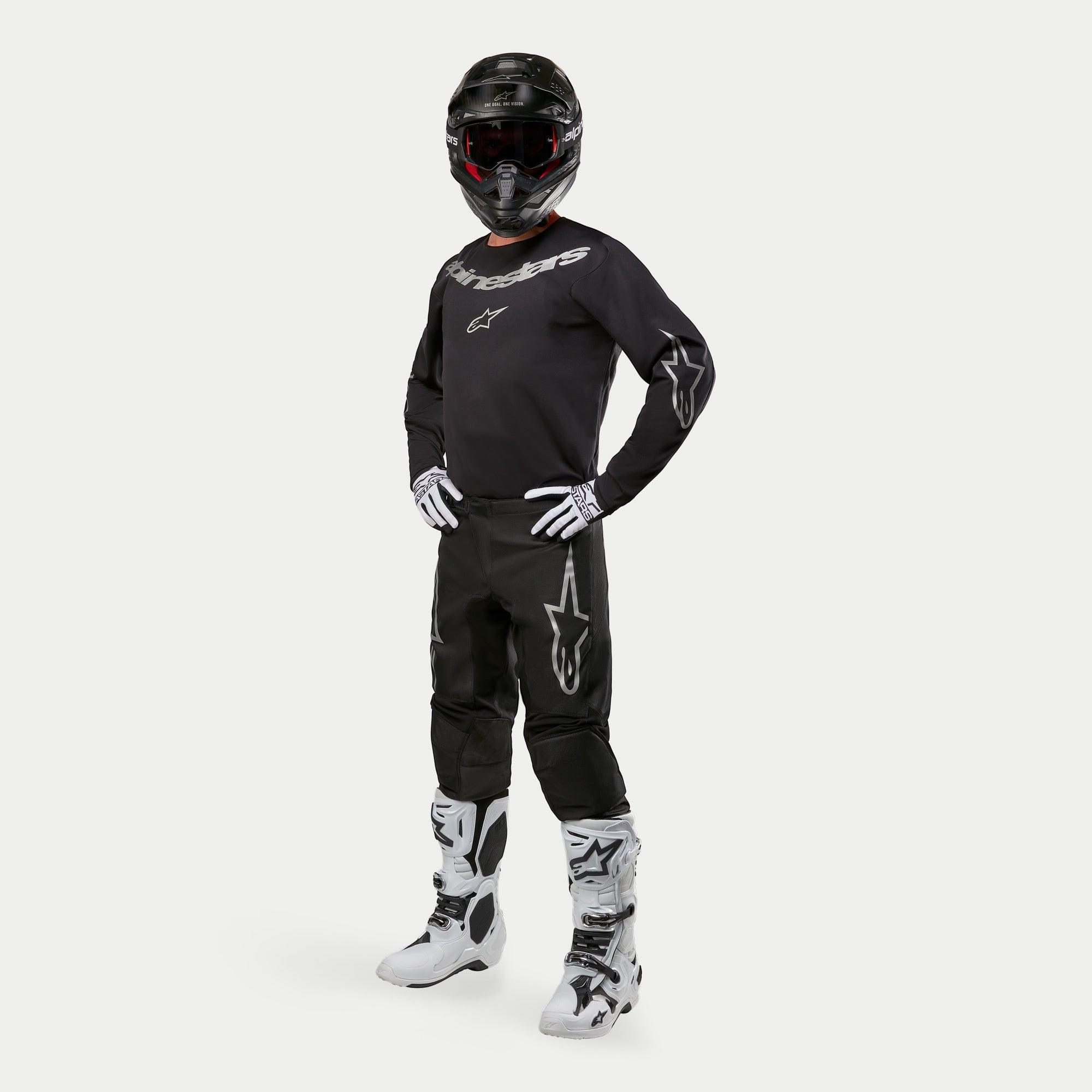 Pilota di motocross con maglia Alpinestars 2024 Fluid Graphite in nero e argento, pantaloni neri coordinati con loghi a stella argento, stivali e guanti bianchi e neri, e casco nero con occhiali rossi, abbigliamento completo da gara off-road