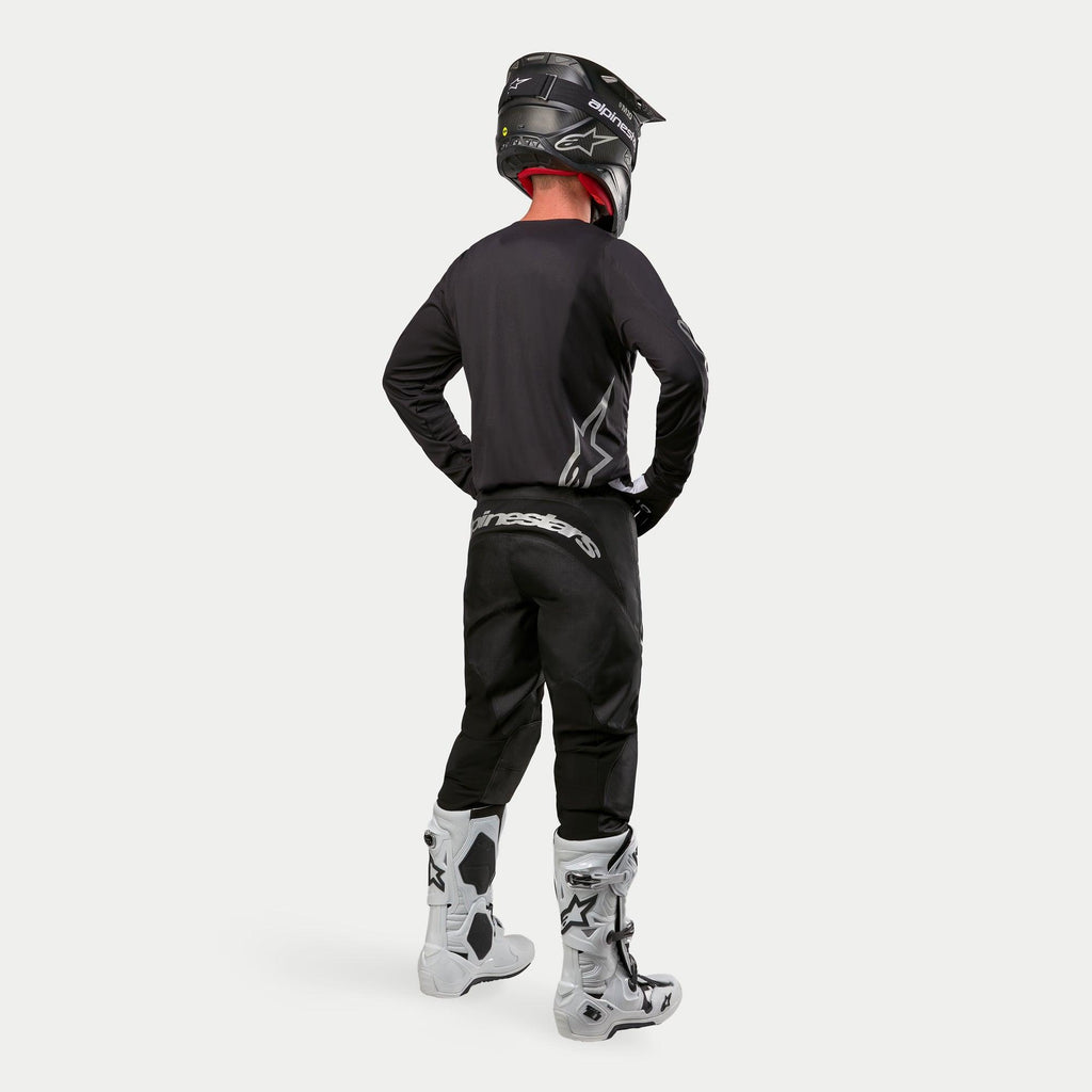 Pilota di motocross con maglia Alpinestars 2024 Fluid Graphite in Black Silver, vista posteriore, maglia a maniche lunghe dal taglio atletico abbinata a pantaloni da motocross neri, casco nero e stivali bianchi e neri, equipaggiamento professionale completo per fuoristrada