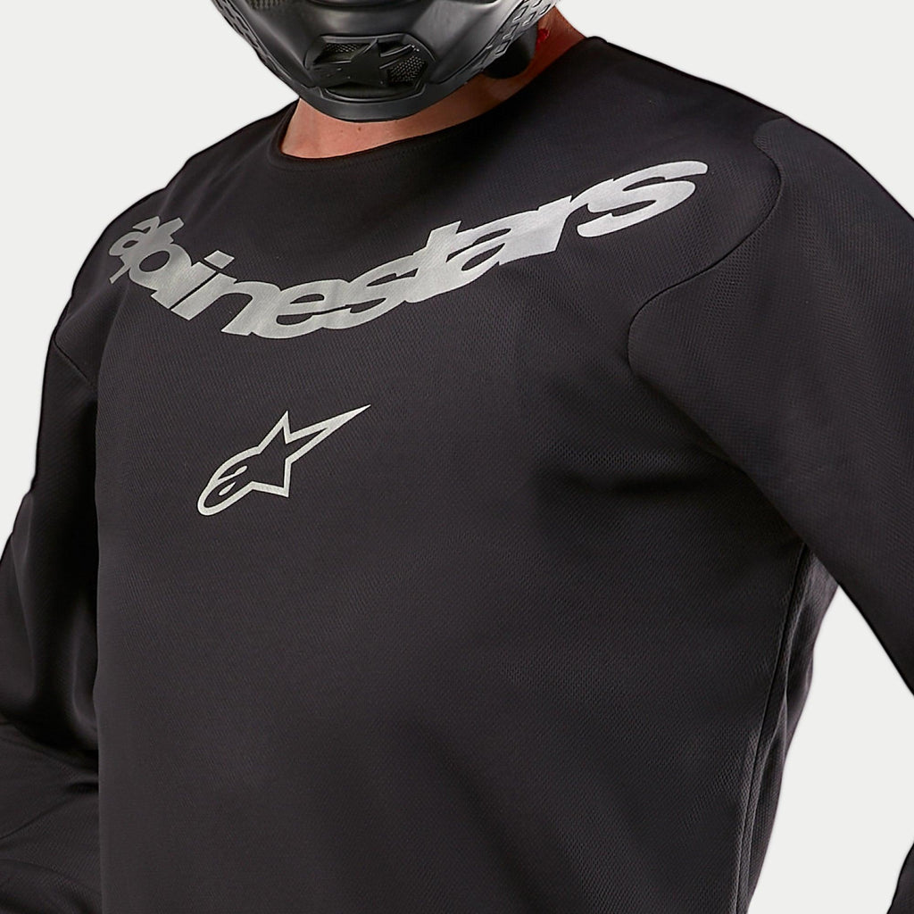 Alpinestars 2024 Fluid Graphite Jersey, maglia da motocross, nero e argento, dettaglio ravvicinato della zona del petto con marchio Alpinestars e logo a stella argento, costruzione in poli-tessuto traspirante, indossata da un pilota con casco nero