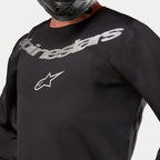 Alpinestars 2024 Fluid Graphite Jersey, maglia da motocross, nero e argento, dettaglio ravvicinato della zona del petto con marchio Alpinestars e logo a stella argento, costruzione in poli-tessuto traspirante, indossata da un pilota con casco nero