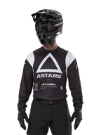 Alpinestars Techdura Jersey, nero, con grande grafica a triangolo bianco sul petto e logo ASTARS, pannelli bianchi sulle spalle, indossata con casco da motocross nero, occhiali e attrezzatura off-road nera coordinata