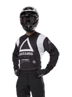 Alpinestars Techdura Jersey, maglia da motocross, nera e bianca, indossata con casco e guanti abbinati, posa con mani sui fianchi su sfondo bianco, con grande logo a triangolo 'A' e marchio ASTARS