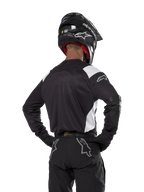 Alpinestars Techdura Jersey, nera con accenti bianchi sulle spalle, vista posteriore di un pilota di motocross con pantaloni e casco neri coordinati, toppe rinforzate sui gomiti, abbigliamento per enduro e gare fuoristrada