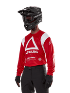 Alpinestars Techdura Jersey, maglia da motocross, rosso brillante e bianco, con un audace design a triangolo bianco e marchio ASTARS, indossata con casco e guanti neri, abbigliamento da gara off-road resistente e traspirante