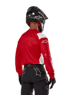 Pilota di motocross visto dal retro che indossa la maglia Alpinestars Techdura in rosso vivo con pannelli bianchi, abbinata a pantaloni da fuoristrada neri, casco nero e guanti neri, abbigliamento coordinato per la guida enduro e motocross.