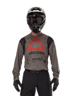 Pilota di motocross con maglia Alpinestars Techdura in Falcon Brown, caratterizzata da un evidente triangolo rosso e logo 'ASTARS' sul petto, abbinata a pantaloni neri, casco nero con occhiali e guanti neri, abbigliamento completo da enduro