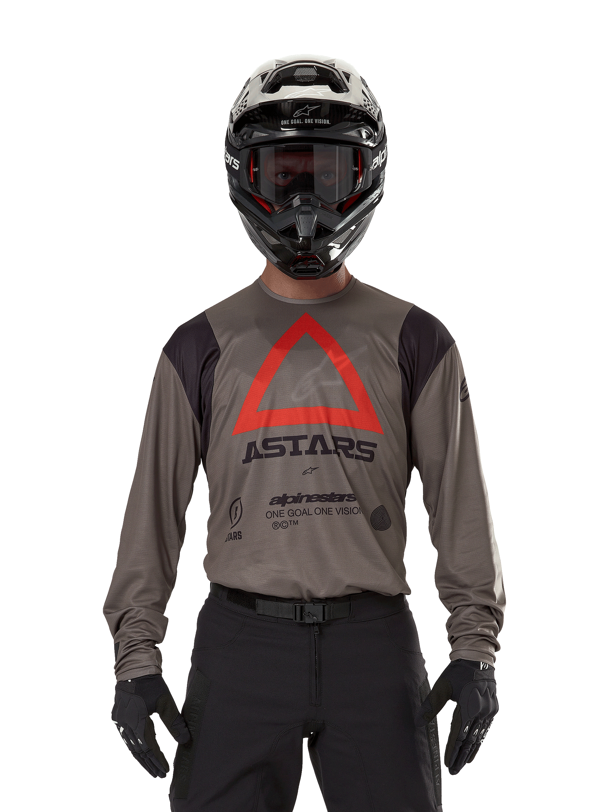 Pilota di motocross con maglia Alpinestars Techdura in Falcon Brown, caratterizzata da un evidente triangolo rosso e logo 'ASTARS' sul petto, abbinata a pantaloni neri, casco nero con occhiali e guanti neri, abbigliamento completo da enduro