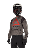 Alpinestars Techdura Jersey, maglia da motocross off-road, Falcon Brown con triangolo rosso e logo nero, indossata con pantaloni neri, casco bianco e nero e guanti neri