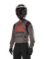Alpinestars Techdura Jersey, maglia da motocross off-road, Falcon Brown con triangolo rosso e logo nero, indossata con pantaloni neri, casco bianco e nero e guanti neri