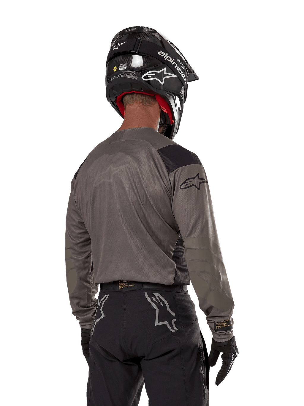 Alpinestars Techdura Jersey, maglia da motocross vista da dietro in Falcon Brown, abbinata a pantaloni off-road e casco neri, con design grafico moderno e profilo posteriore allungato per l'enduro