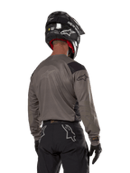 Alpinestars Techdura Jersey, maglia da motocross vista da dietro in Falcon Brown, abbinata a pantaloni off-road e casco neri, con design grafico moderno e profilo posteriore allungato per l'enduro