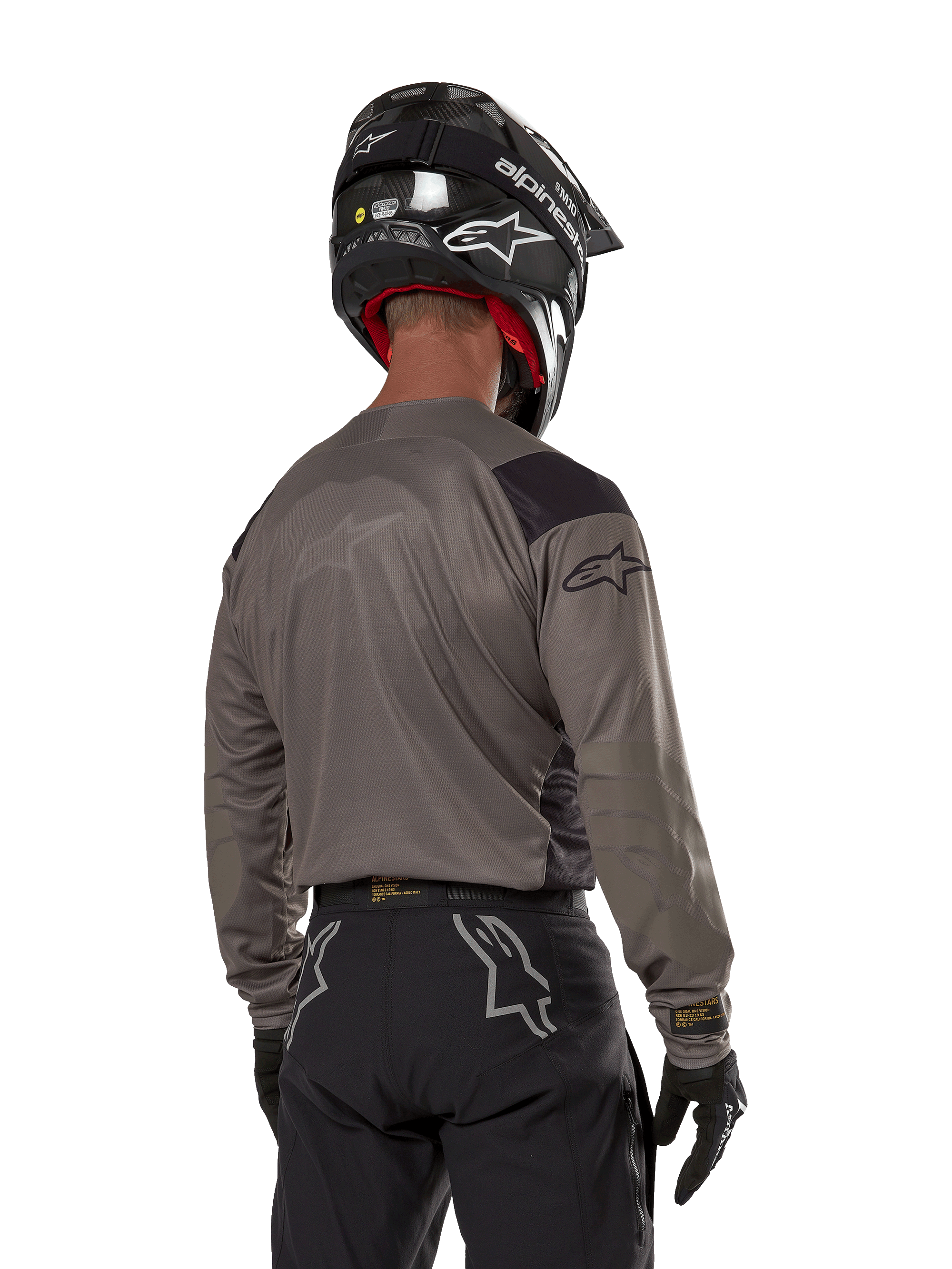 Alpinestars Techdura Jersey, maglia da motocross vista da dietro in Falcon Brown, abbinata a pantaloni off-road e casco neri, con design grafico moderno e profilo posteriore allungato per l'enduro
