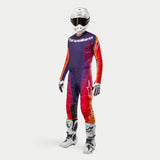 Abbigliamento completo da motocross Alpinestars Techstar Pneuma 2024, casco bianco con occhiali neri, maglia e pantaloni viola, arancione e blu, guanti e stivali bianchi con dettagli neri.