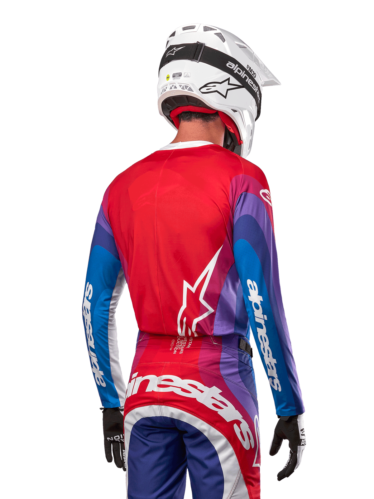 Pilota di motocross visto da dietro con maglia Alpinestars Techstar Pneuma 2024 in Blu Mars Red White, caratterizzata da un retro rosso con logo a stella bianco, maniche blu e viola con branding bianco, coordinata con pantaloni abbinati e un casco bianco per gare fuoristrada.