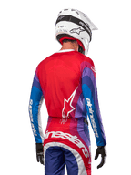 Pilota di motocross visto da dietro con maglia Alpinestars Techstar Pneuma 2024 in Blu Mars Red White, caratterizzata da un retro rosso con logo a stella bianco, maniche blu e viola con branding bianco, coordinata con pantaloni abbinati e un casco bianco per gare fuoristrada.