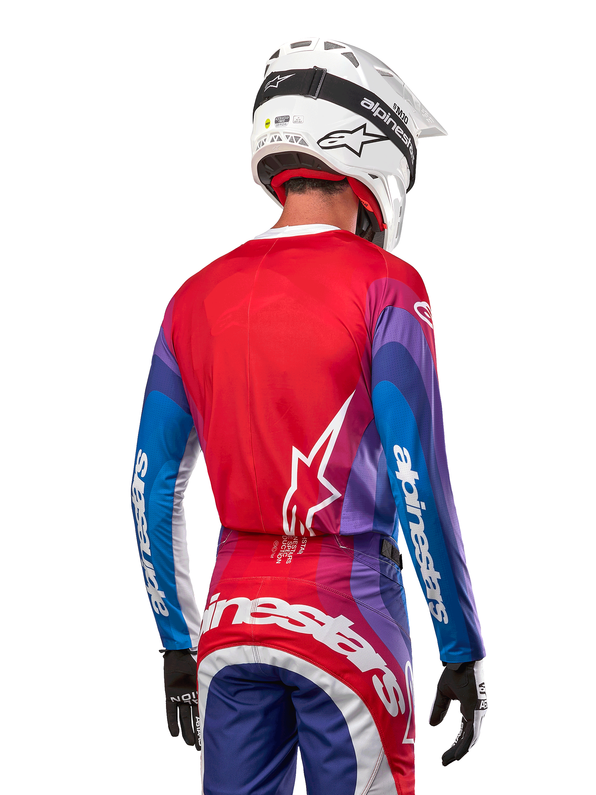 Pilota di motocross visto da dietro con maglia Alpinestars Techstar Pneuma 2024 in Blu Mars Red White, caratterizzata da un retro rosso con logo a stella bianco, maniche blu e viola con branding bianco, coordinata con pantaloni abbinati e un casco bianco per gare fuoristrada.