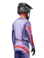 Alpinestars Techstar Ocuri Jersey 2024, maglia da motocross, viola e arancione acceso, vista posteriore su un pilota con equipaggiamento coordinato e casco nero, con grandi scritte Alpinestars arancioni sulle maniche e sulla parte bassa della schiena