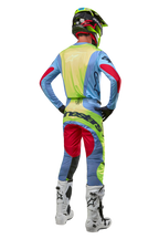Pilota di motocross visto di spalle con maglia Alpinestars Techstar Ocuri 2024, celeste con accenti giallo fluo e rosso berry, pantaloni da gara coordinati, stivali bianchi e casco neon, equipaggiamento completo da fuoristrada per competizioni professionali