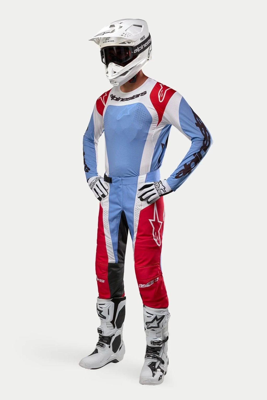 Pilota di motocross con equipaggiamento completo Alpinestars: maglia Techstar Ocuri 2024 in azzurro, rosso e bianco, pantaloni abbinati, casco bianco con occhiali, stivali bianchi e guanti bianchi e neri.