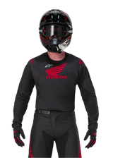 Pilota di motocross con abbigliamento completo da gara, maglia Honda Racer Iconic nera con logo ad ala rosso, pantaloni neri abbinati con marchio Honda rosso, casco nero con occhiali e guanti neri e rossi, completo coordinato per corse fuoristrada