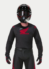 Pilota di motocross con maglia Alpinestars Honda Racer Iconic nero e rosso, grande logo Honda ad ala rosso sul petto e logo Astar bianco, abbinata a casco, occhiali e guanti neri per un kit completo da gara fuoristrada