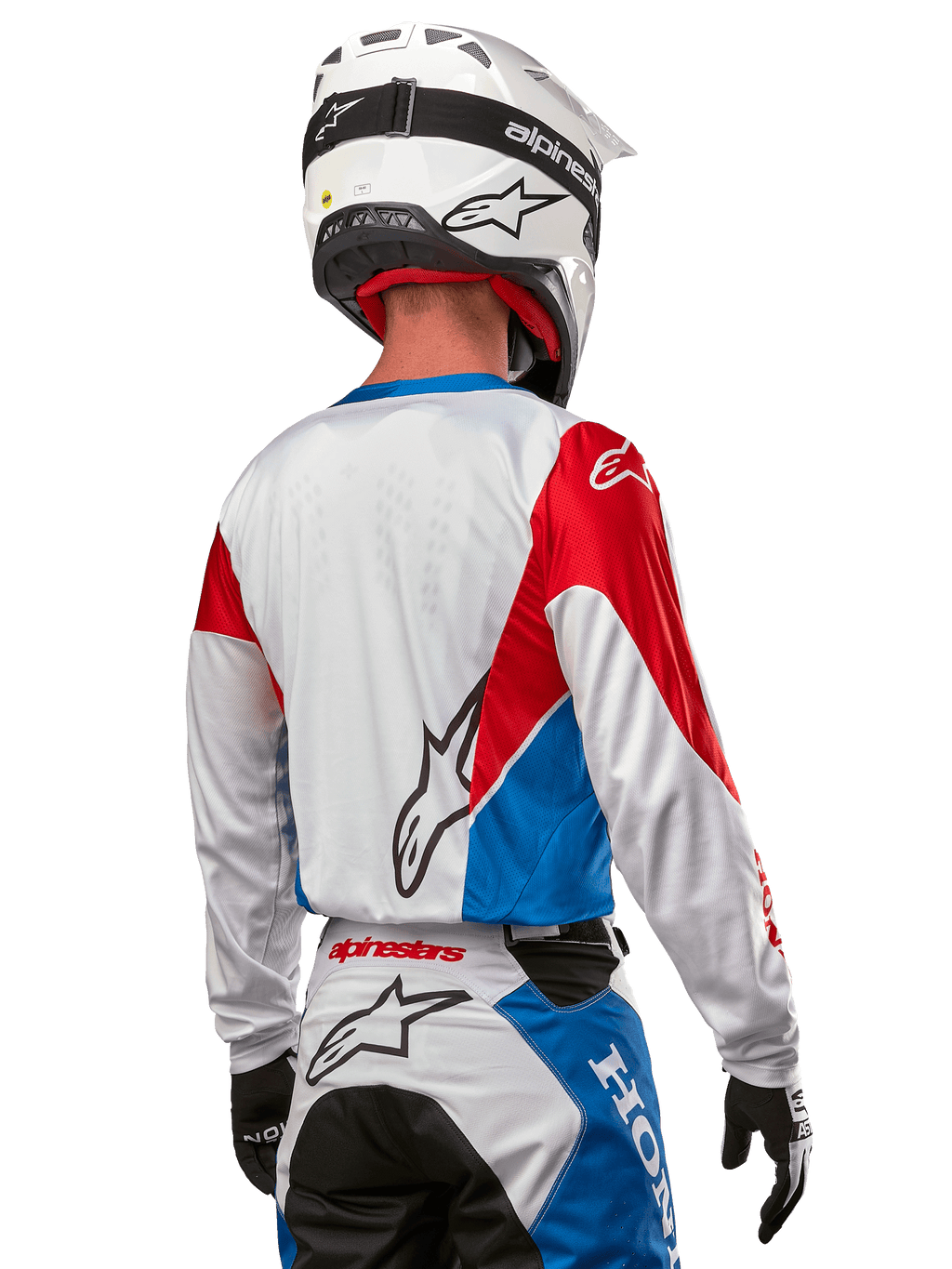 Pilota di motocross con maglia Alpinestars Honda Racer Iconic, vista posteriore, bianco con pannelli blu e rosso brillante, con logo Alpinestars sul retro, abbinata a casco bianco e pantaloni da motocross coordinati.