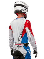 Pilota di motocross con maglia Alpinestars Honda Racer Iconic, vista posteriore, bianco con pannelli blu e rosso brillante, con logo Alpinestars sul retro, abbinata a casco bianco e pantaloni da motocross coordinati.