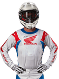 Alpinestars Honda Racer Iconic Jersey, maglia da motocross bianca con logo Honda rosso e accenti blu e rossi vivaci, indossata da un pilota con casco e guanti bianchi, abbigliamento tecnico per competizioni motocross e fuoristrada.