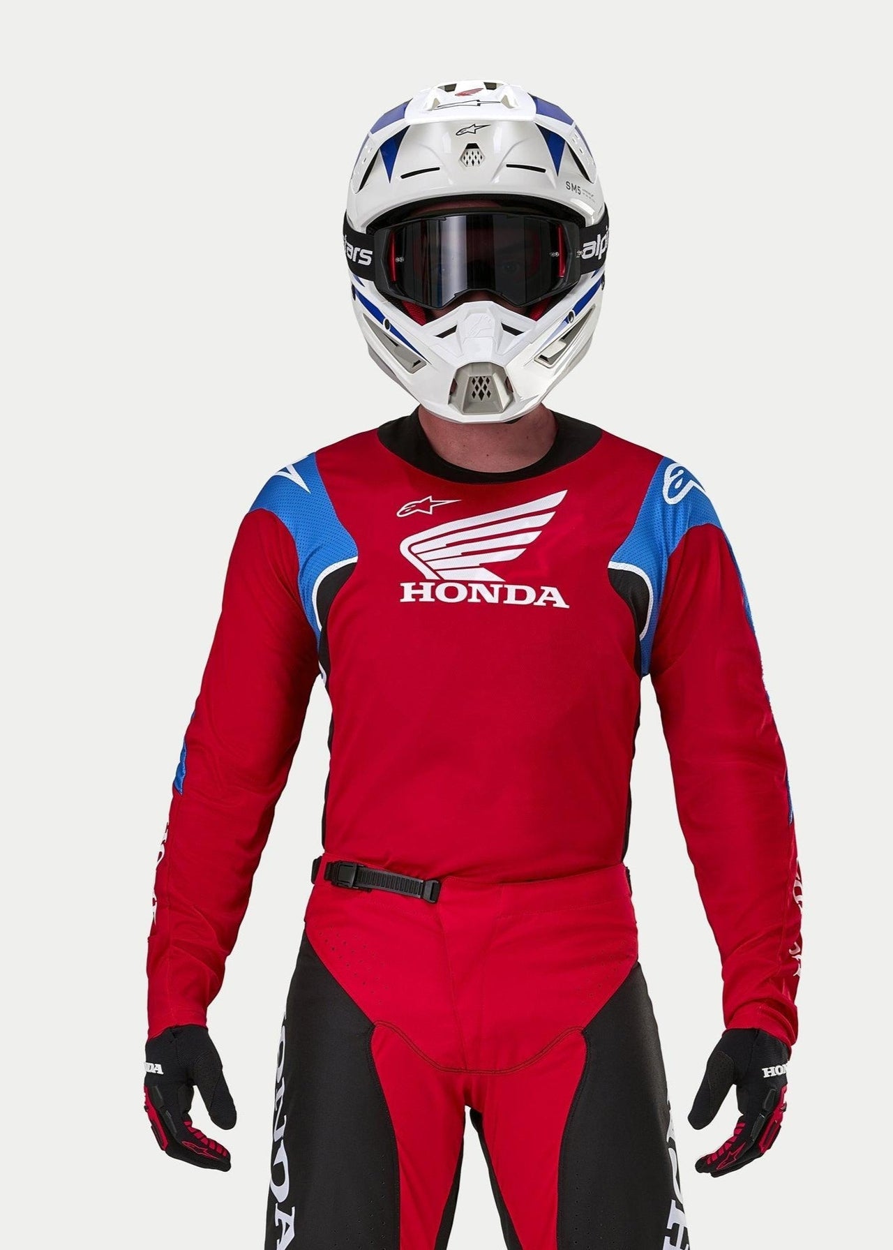 Pilota di motocross che indossa la maglia Alpinestars Honda Racer Iconic, rosso acceso con accenti blu e bianchi e marchio Honda, abbinata a un casco bianco e blu, occhiali scuri e pantaloni da motocross coordinati rossi e neri, abbigliamento tecnico per il fuoristrada
