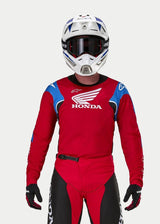 Pilota di motocross che indossa la maglia Alpinestars Honda Racer Iconic, rosso acceso con accenti blu e bianchi e marchio Honda, abbinata a un casco bianco e blu, occhiali scuri e pantaloni da motocross coordinati rossi e neri, abbigliamento tecnico per il fuoristrada