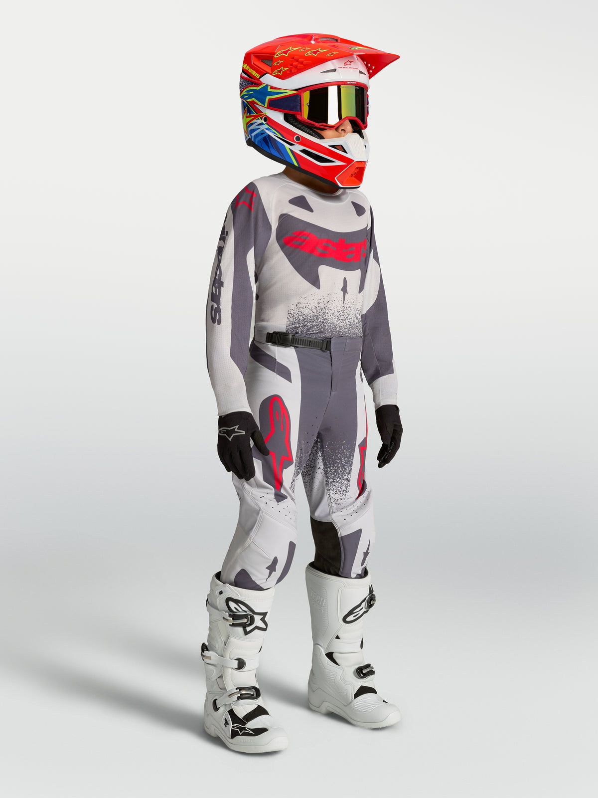 Ragazzo Supertech Swan Gear Set e maglia Alpinestars, completo motocross grigio e rosso fluo con stivali da corsa bianchi.