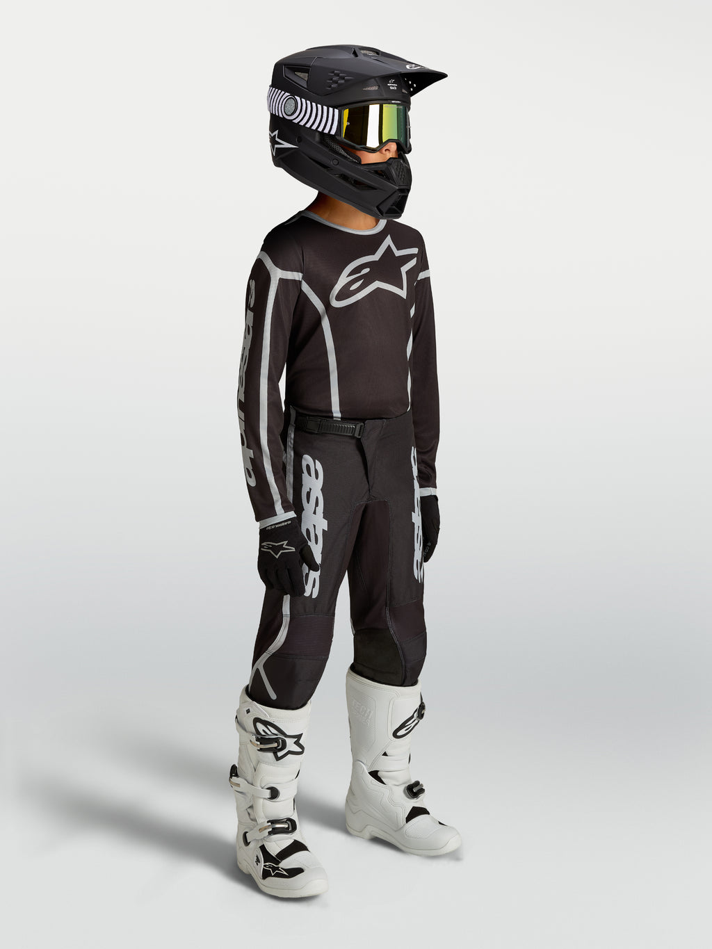 Alpinestars maglia e completo motocross Fluid Apex V2 ragazzo, neri e argento, traspiranti, con casco e stivali bianchi.