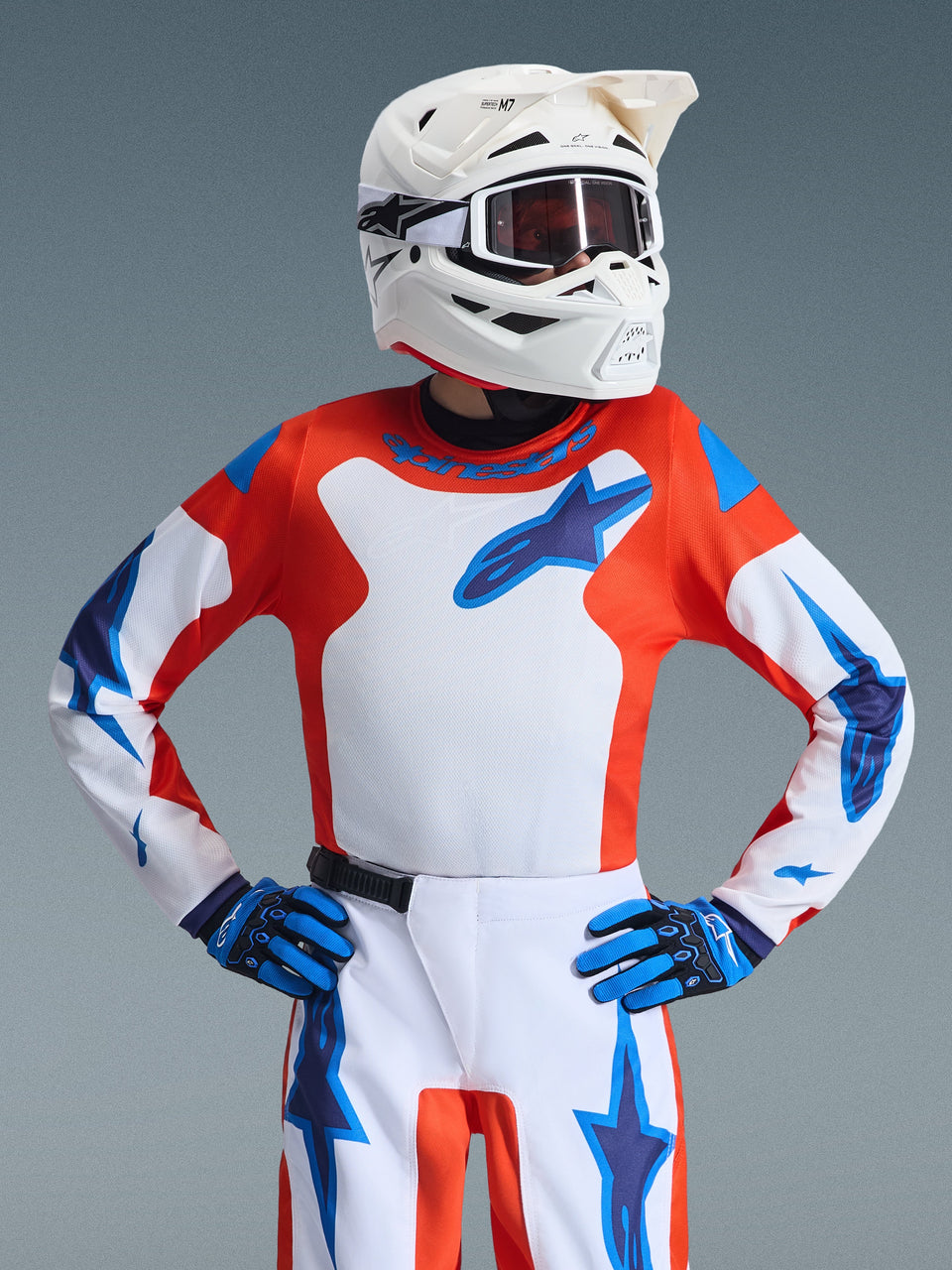 Giovane pilota di motocross con maglia Alpinestars 2026 Youth Fluid Grid, arancione e blu UCLA con pannello centrale bianco e logo blu, abbinata a pantaloni coordinati, guanti blu e neri e casco bianco con occhiali, set completo di abbigliamento da corsa per fuoristrada.