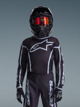 Giovane pilota di motocross con maglia Alpinestars Youth Fluid Apex 2026 in nero e grigio, pantaloni coordinati, casco nero con occhiali riflettenti rossi e guanti neri, abbigliamento da corsa fuoristrada coordinato