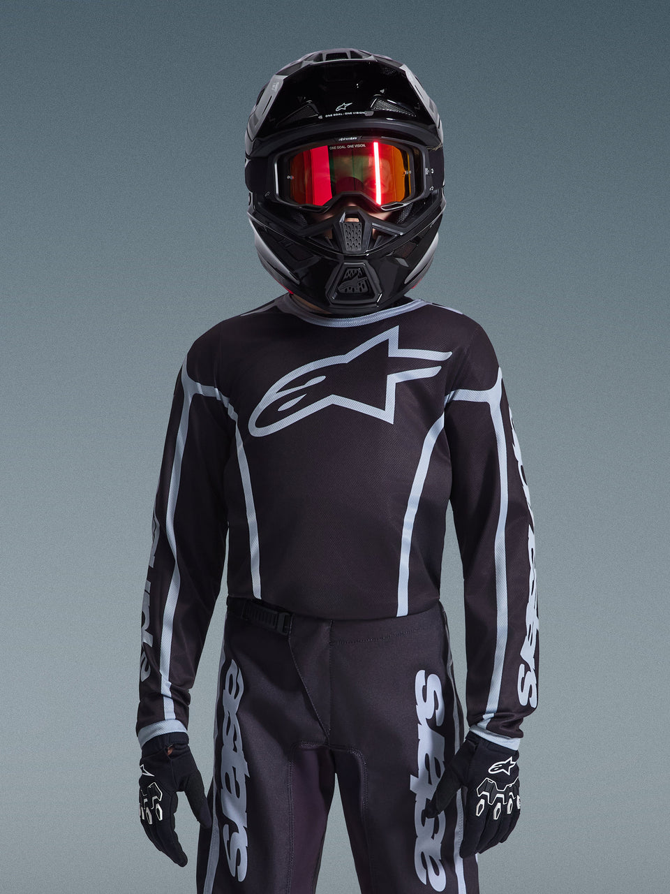 Giovane pilota di motocross con maglia Alpinestars Youth Fluid Apex 2026 in nero e grigio, pantaloni coordinati, casco nero con occhiali riflettenti rossi e guanti neri, abbigliamento da corsa fuoristrada coordinato
