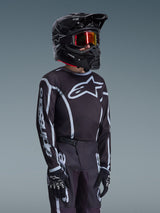 Alpinestars Maglia Youth Fluid Apex 2026 in nero e grigio, indossata da un giovane pilota con casco nero coordinato e occhiali con lenti a specchio, con logo Alpinestars evidente sul petto e sulle maniche, parte di un set di abbigliamento da motocross coordinato.