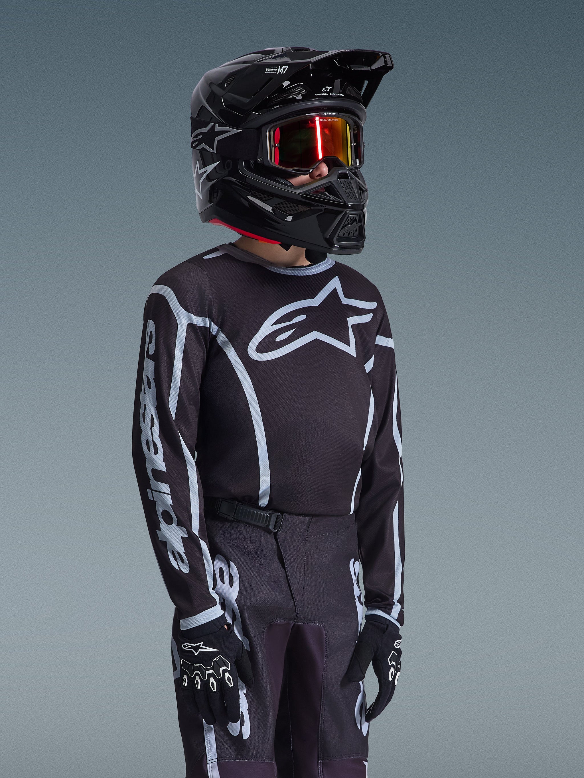 Alpinestars Maglia Youth Fluid Apex 2026 in nero e grigio, indossata da un giovane pilota con casco nero coordinato e occhiali con lenti a specchio, con logo Alpinestars evidente sul petto e sulle maniche, parte di un set di abbigliamento da motocross coordinato.