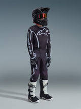 Giovane pilota di motocross con maglia Alpinestars Youth Fluid Apex 2026 in nero e grigio, pantaloni coordinati neri e grigi con grafiche bianche, casco nero con occhiali dalle lenti rosse, guanti neri e stivali da motocross bianchi e neri, kit completo da gara fuoristrada.