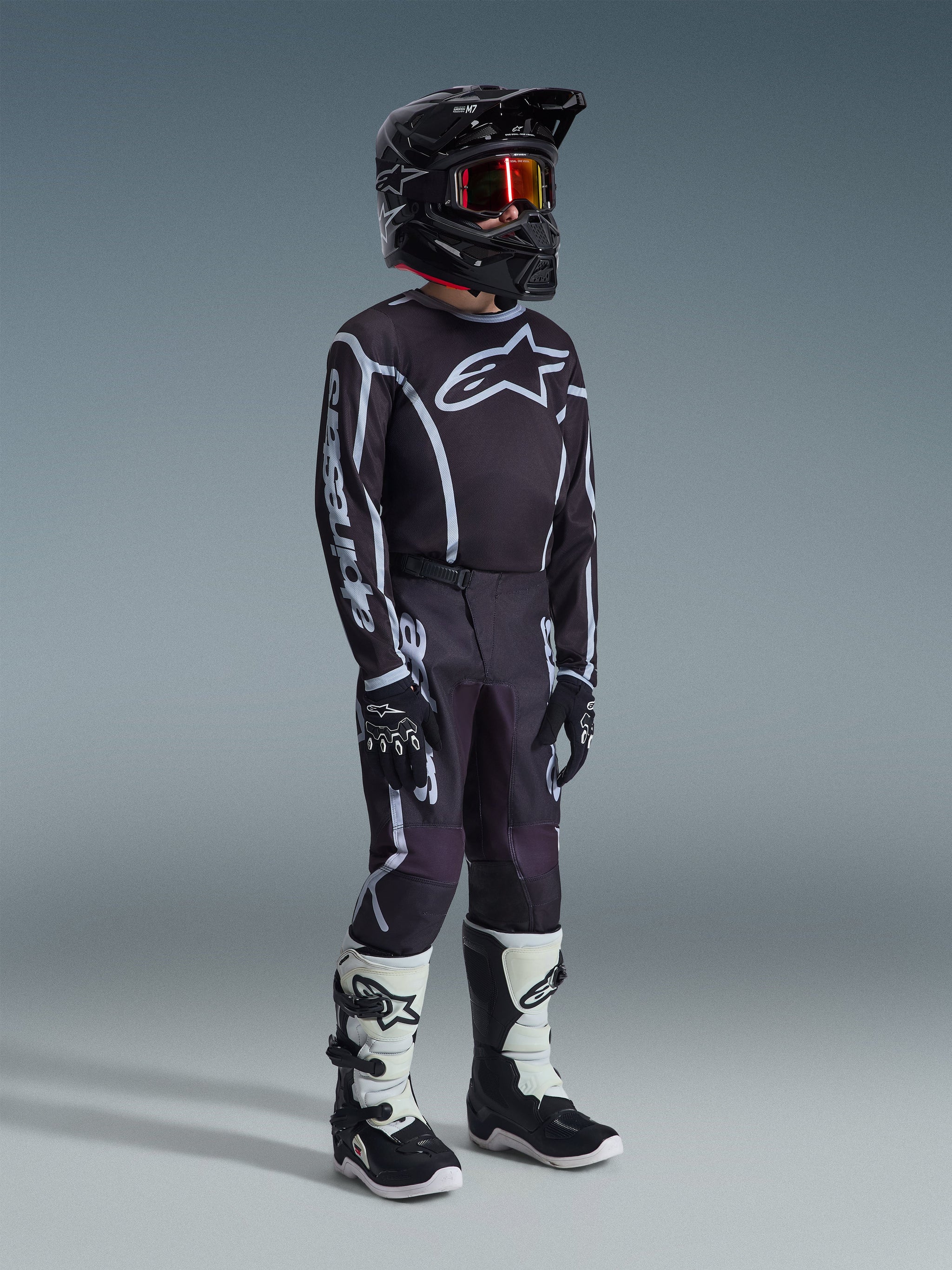 Giovane pilota di motocross con maglia Alpinestars Youth Fluid Apex 2026 in nero e grigio, pantaloni coordinati neri e grigi con grafiche bianche, casco nero con occhiali dalle lenti rosse, guanti neri e stivali da motocross bianchi e neri, kit completo da gara fuoristrada.