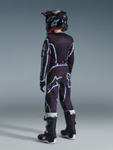 Maglia Alpinestars Youth Fluid Apex 2026, maglia da motocross, nero e grigio, indossata con pantaloni Youth Fluid Grid coordinati, casco nero e stivali bianchi e neri, vista posteriore dell'attrezzatura completa da off-road per giovani piloti