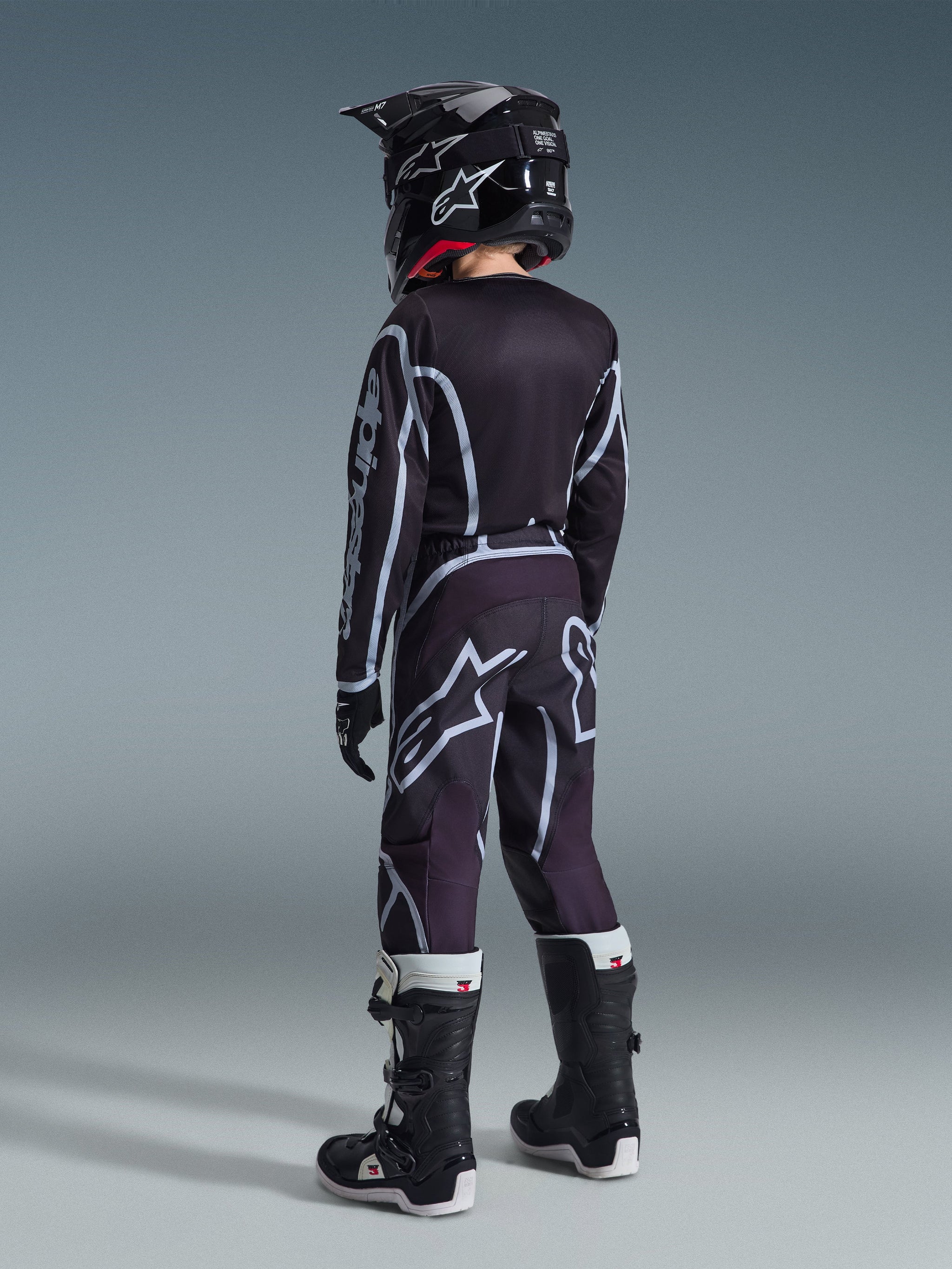 Maglia Alpinestars Youth Fluid Apex 2026, maglia da motocross, nero e grigio, indossata con pantaloni Youth Fluid Grid coordinati, casco nero e stivali bianchi e neri, vista posteriore dell'attrezzatura completa da off-road per giovani piloti