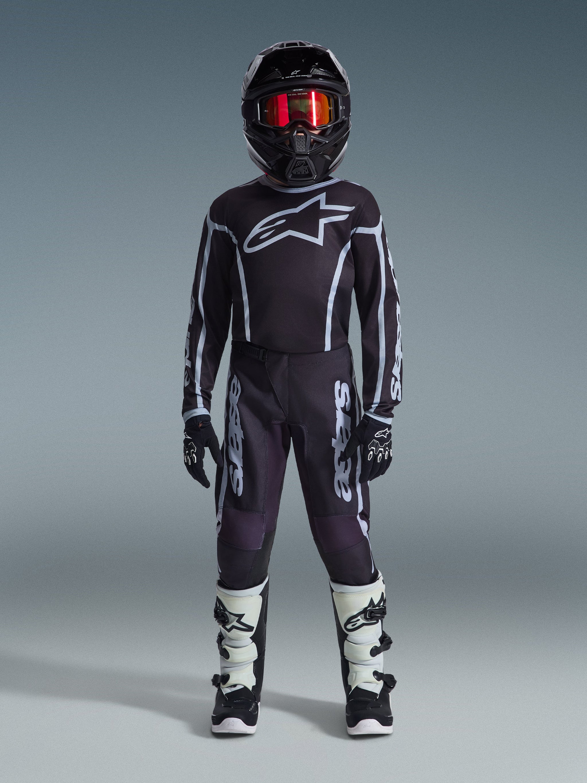 Giovane pilota di motocross con maglia Alpinestars 2026 Youth Fluid Apex in nero e grigio, pantaloni coordinati, casco nero con occhiali iridescenti rossi, guanti bianchi e neri e stivali da motocross, set completo di abbigliamento per corse fuoristrada per ragazzi