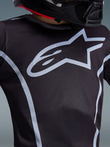 Alpinestars Youth Fluid Apex Jersey 2026, primo piano del design sul petto, nero e grigio, con un prominente logo a stella grigio chiaro e profili sportivi, indossata con un casco nero, abbigliamento tecnico da motocross per giovani piloti