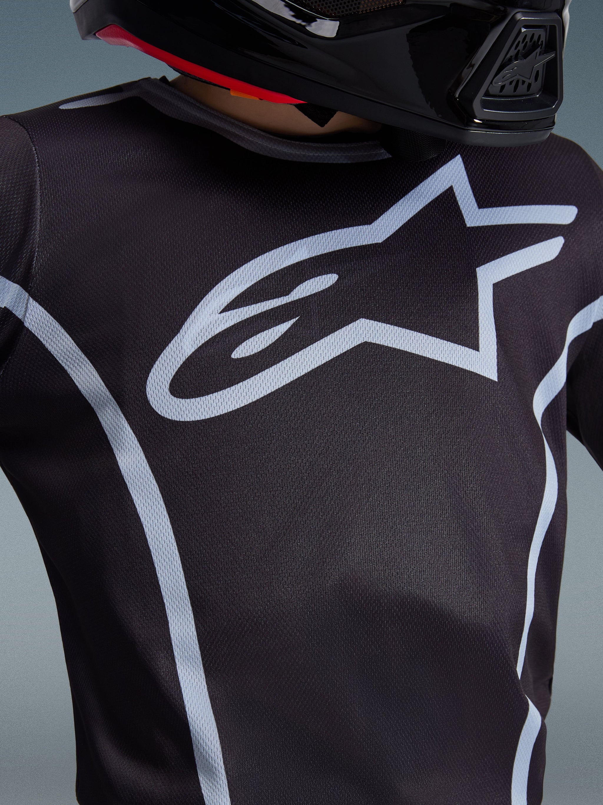 Alpinestars Youth Fluid Apex Jersey 2026, primo piano del design sul petto, nero e grigio, con un prominente logo a stella grigio chiaro e profili sportivi, indossata con un casco nero, abbigliamento tecnico da motocross per giovani piloti