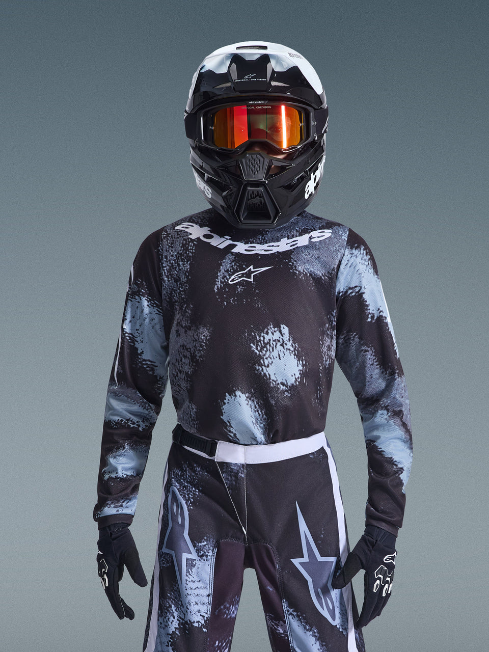 Alpinestars 2026 Youth Fluid Lahnd Jersey, maglia da motocross, Iron Camo, indossata da un giovane pilota con pantaloni coordinati, casco bianco e nero con maschera arancione e guanti neri, caratterizzata da un design grigio e nero effetto schizzato con loghi bianchi, abbigliamento completo da fuoristrada per giovani piloti.