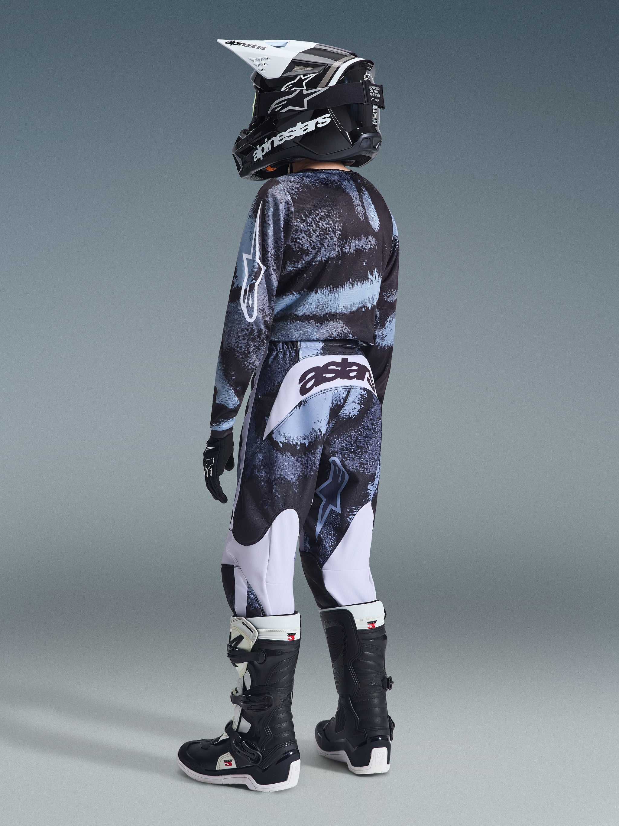 Giovane pilota di motocross che indossa la maglia Alpinestars Youth Fluid Lahnd 2026 e i pantaloni coordinati, motivo mimetico nero e grigio con accenti azzurri, marchio Astars bianco, casco nero con dettagli bianchi, stivali neri e bianchi, completo da corsa fuoristrada coordinato