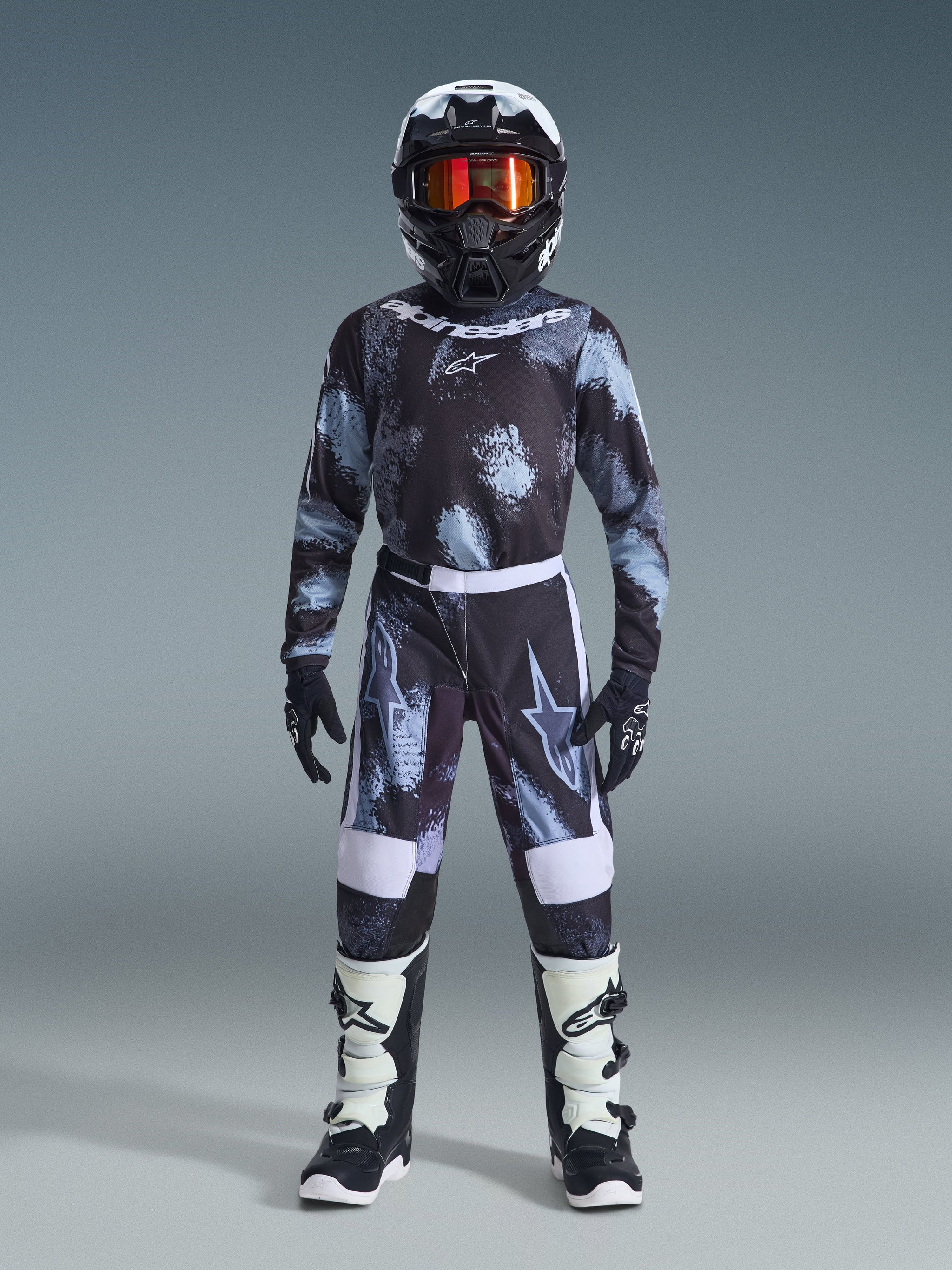 Giovane pilota di motocross con abbigliamento completo da gara, maglia Alpinestars Youth Fluid Lahnd 2026 in Iron Camo con motivo nero, grigio e azzurro, pantaloni abbinati, casco bianco e nero con maschera dalle lenti arancioni, stivali bianchi e neri, set di equipaggiamento coordinato per la guida fuoristrada