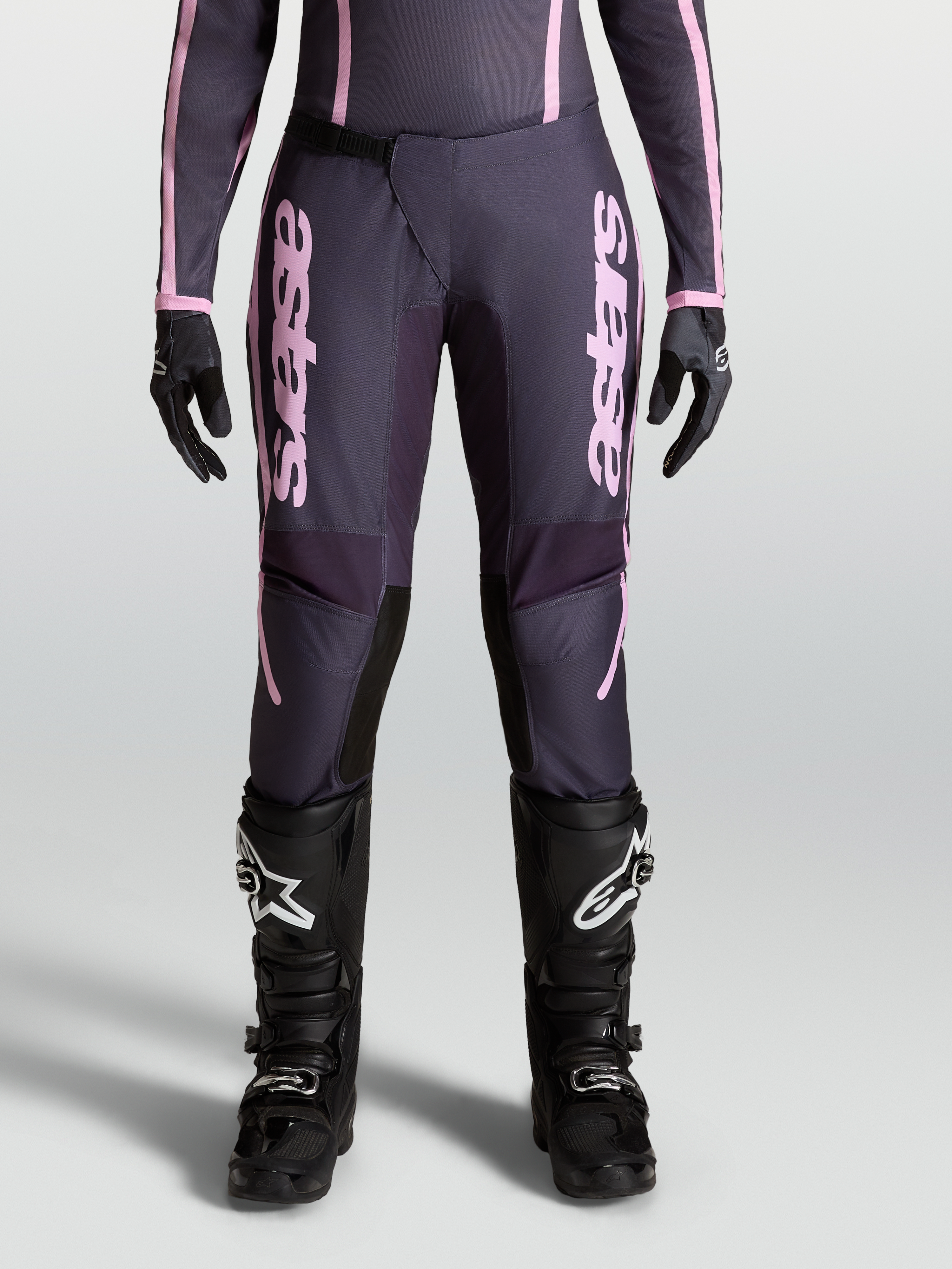 Alpinestars Pantaloni Stella Fluid Apex V2, pantaloni motocross da donna, grigio e rosa, chiusura a cricchetto e design precurvato, indossati con stivali neri per fuoristrada.