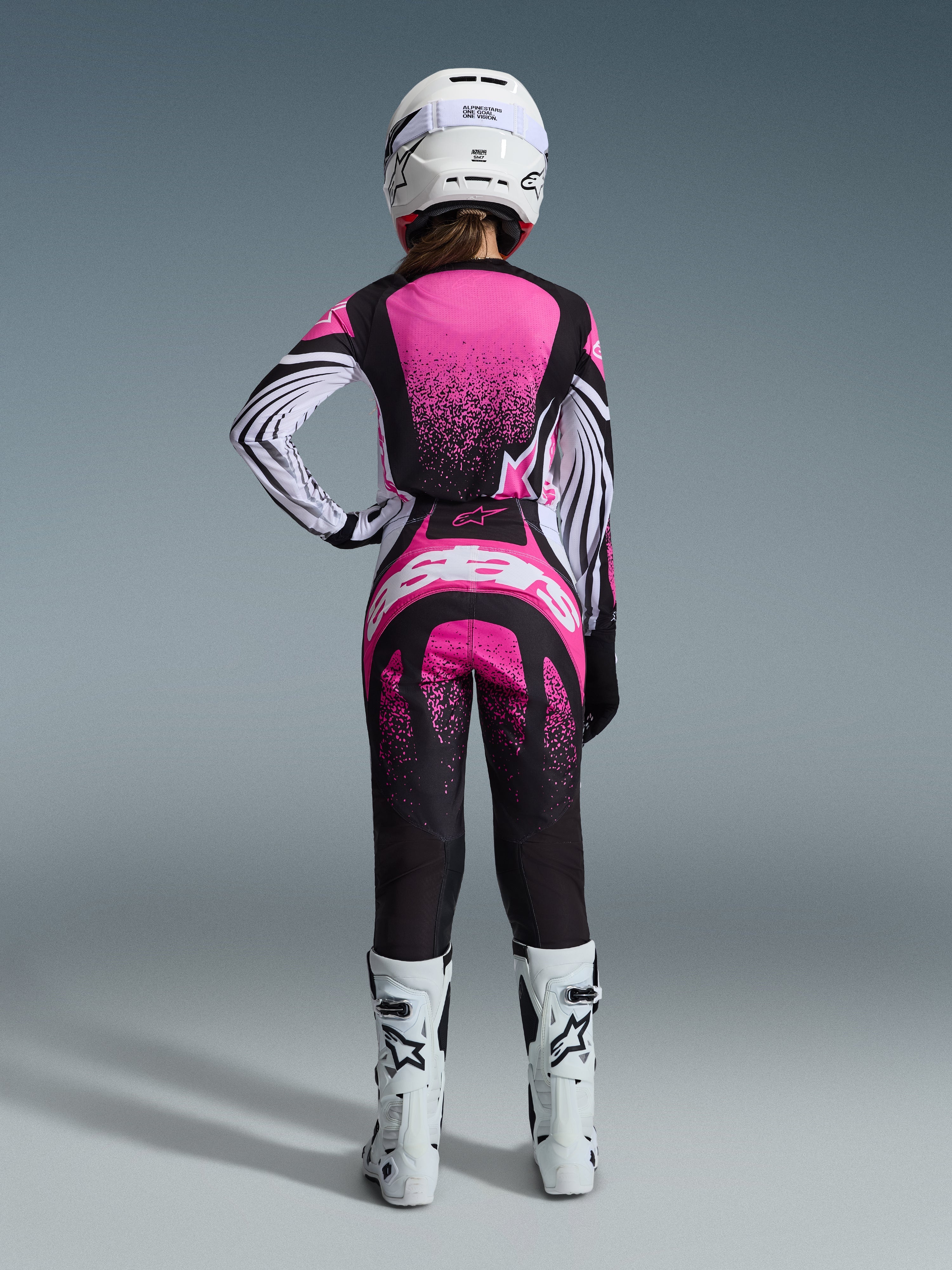 Alpinestars 2026 Stella Techstar Nomur Gear Set, pilota di motocross donna vista da dietro, maglia rosa e nera con maniche a righe bianche e motivo puntinato, pantaloni coordinati con logo Alpinestars bianco, casco bianco e stivali off-road bianchi