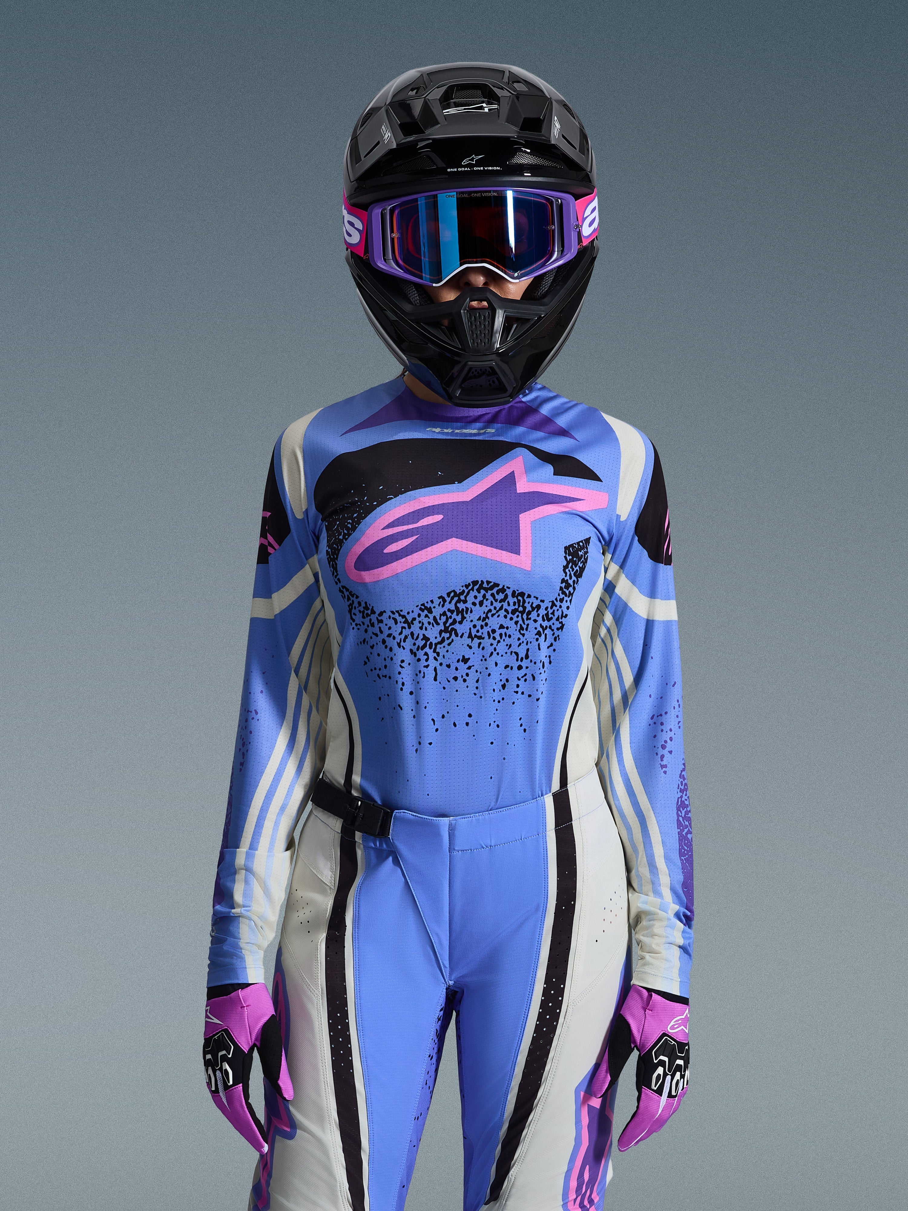 Pilota di motocross donna che indossa il completo Alpinestars 2026 Stella Techstar Nomur, maglia blu con logo a stella rosa e viola, pantaloni blu coordinati con pannelli laterali bianchi e neri, casco nero con occhiali viola e blu e guanti rosa e neri, kit completo da gara off-road da donna